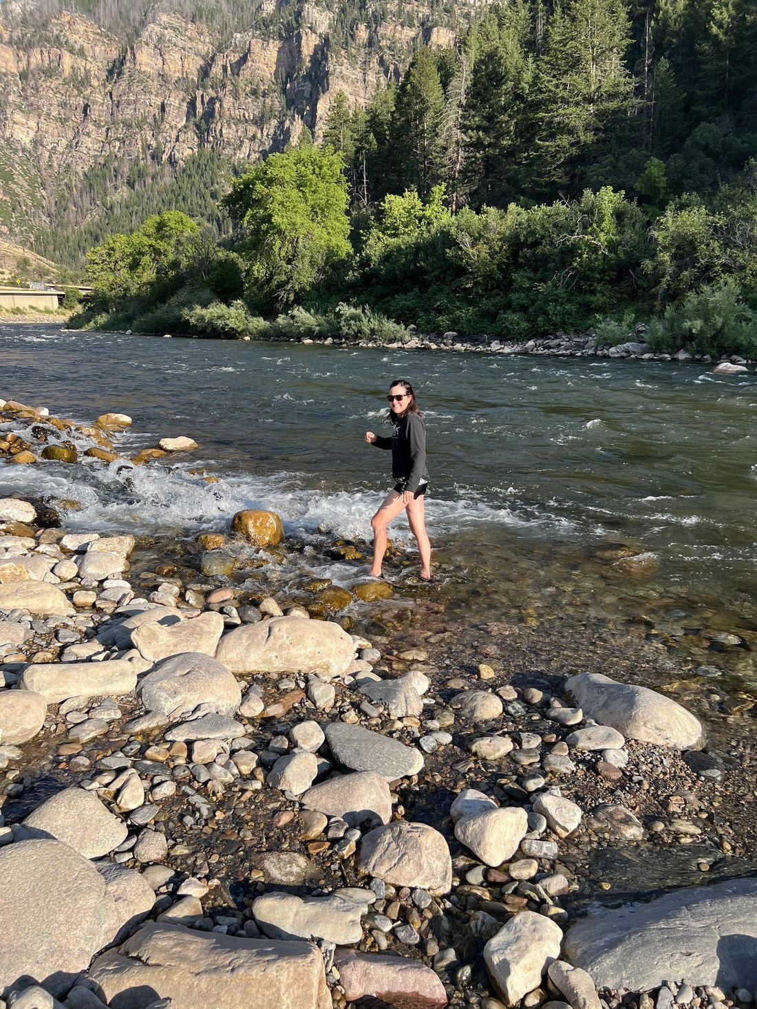 Suzanne wading in a river in a mountain canyon, smiling, dressed casually outdoors.